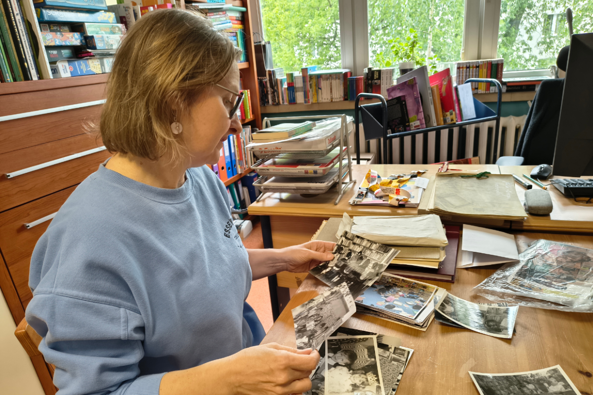 Kobieta w niebieskim swetrze i okularach siedzi przy drewnianym biurku w pokoju z książkami, oglądając stare czarno-białe fotografie rozłożone na stole, w tle regały z książkami, dokumenty i okno z widokiem na drzewa.